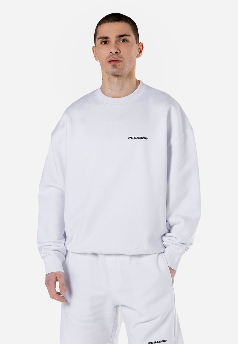 Sudadera blanca de tela suave con mangas largas y puños acanalados, que presenta un pequeño logo negro "PEGADOR" en el frontal.