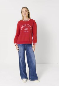 Felpa rossa con testo bianco, dotata di scollatura rotonda e polsini a righe, abbinata a jeans larghi blu e scarpe bianche.