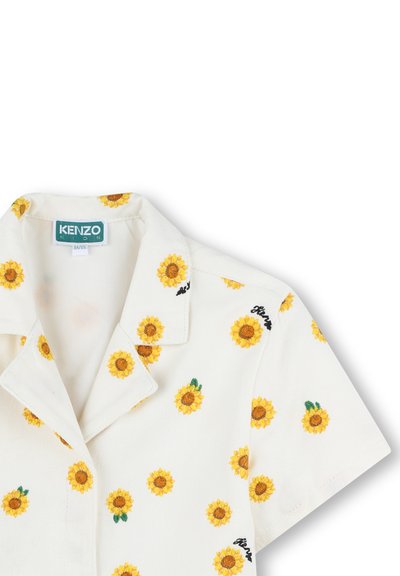 Chemise blanche à manches courtes avec des tournesols jaunes brodés et le texte noir "floral", dotée d'un col en V et d'une texture douce. Étiquette KENZO visible.
