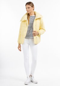 Geel gewatteerde jas met een hoge kraag, knoopsluiting en elastische manchetten, gedragen over een gestreept shirt en gecombineerd met witte skinny jeans en sneakers.
