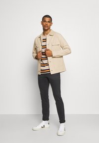 Chaqueta de algodón beige con dos bolsillos frontales, usada sobre una camisa de rayas en tonos de azul y naranja, combinada con jeans oscuros y zapatillas blancas.
