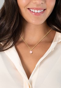 Collana in oro a catena con un pendente rotondo di pietra rosa, indossata sopra una blusa beige chiaro con dettagli a bottoni.