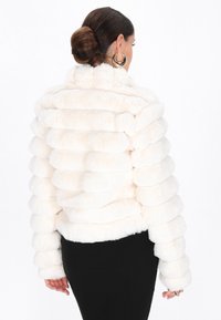Kvinde med mørkt hår i en knold, iført store guld hoopsøreringe og en hvid tekstureret pelsjakke, der vender bort, parret med en sort nederdel.