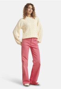 Chunky cream cable-knit sweater with voluminous sleeves paired with high-waisted, flared pink pants. Model stands against a white background.