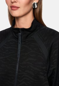 Schwarze Jacke mit Reißverschluss und strukturierter Musterung, ausgestattet mit einem hohen Kragen und dezentem Glanz. Silberne quadratische Ohrringe ergänzen das Design.