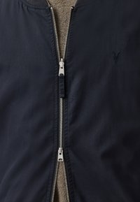 Chaqueta navy con cremallera plateada, cuello acanalado y un logo bordado. La tela parece suave con un ligero brillo.