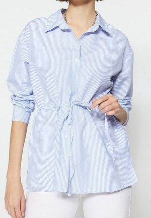 Vrouw draagt een lichtblauwe en witte verticale gestreepte blouse met knopen, met een trekkoord in de taille en een witte broek.