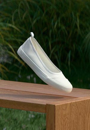 Hvit slip-on canvas-sko svever over et trebord utendørs med grønt gress og planter i bakgrunnen.