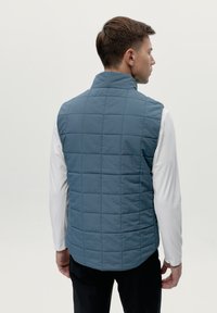Gilet leggero imbottito in una tonalità di blu tenue, con colletto alto e texture liscia, indossato sopra una camicia bianca a maniche lunghe.