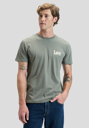 Giovane uomo con capelli ricci che indossa una maglietta verde con logo Lee e jeans blu, in piedi con una mano in tasca su sfondo grigio.