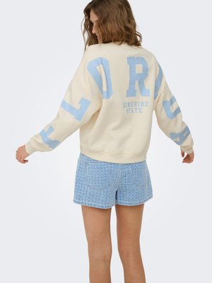 Woman with wavy hair wearing cream sweatshirt with large blue letters and light blue textured shorts, standing with arms slightly extended.