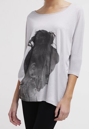 Kobieta w jasnoszarej koszulce z długim rękawem z czarną grafiką sylwetki z kokiem oraz panoramą miasta na przodzie.