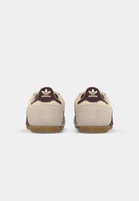 Sneaker in suede beige con accenti bordeaux e rete testurizzata. Presenta suola in gomma e logo Adidas sul tallone. Vista posteriore, design minimalista.