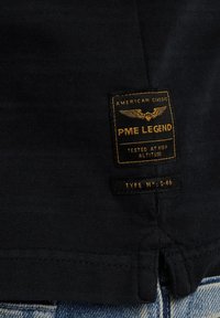 Camiseta de algodón negra con una etiqueta texturizada que presenta un texto dorado y un emblema con alas, que indica la marca y el tipo, cosida en la parte inferior izquierda.