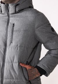 Giubbotto grigio trapuntato con cappuccio, dotato di una cerniera nera, tasche laterali e polsini a coste. Il materiale ha una finitura testurizzata.