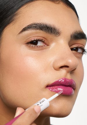 Applicateur de gloss à lèvres tenu près de lèvres pleines, recouvertes d'un gloss rose brillant. Le teint est clair, avec une texture lisse et un maquillage naturel.