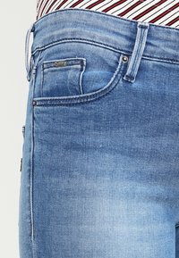 Vaaleansiniset farkut, joissa on klassinen viiden taskun malli, hillitty haalistuma ja metalliset kiinnikkeet ja yksityiskohdat.