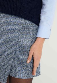 Suéter de punto navy sobre una falda tejida texturizada en azul y negro. Primer plano de una mano descansando en el dobladillo de la falda. Puños de camisa azul claro visibles.