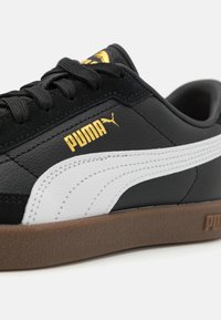 PUMA sneakers med svart skinnfôr, hvit stripe, gulllogo og brun gummisåle. Snørene og teksturerte detaljer er synlige.