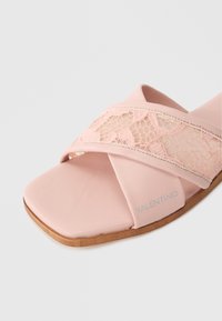 Pink slide sandal featuring a textured lace strap overlay, flat sole, and light brown outsole with branding on the side.