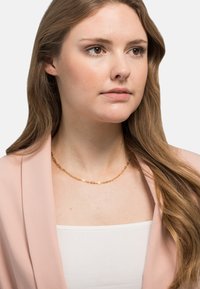 Gull lenke halskjede har avlange ovale og rektangulære lenker. Båret med en lyserosa blazer over en hvit topp. Jevn tekstur.