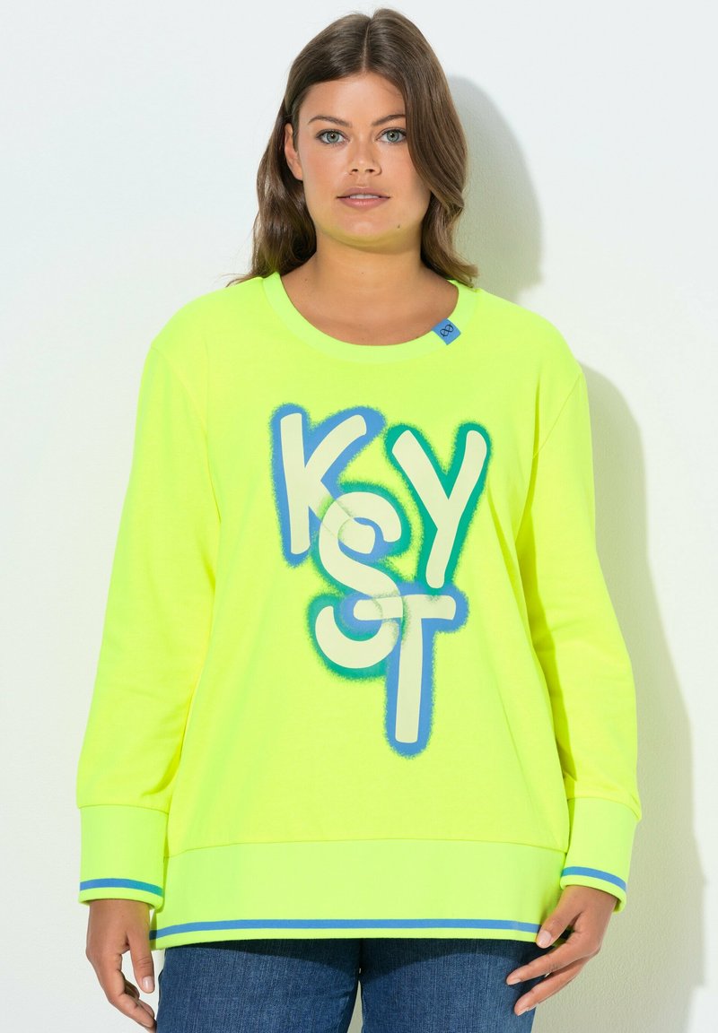 Donna con capelli castani che indossa una felpa giallo neon con lettere "KOST" blu e verdi e polsini a righe blu, abbinata a jeans blu.