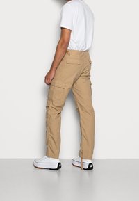 Beżowe spodnie typu cargo z wieloma kieszeniami i luźnym krojem, w połączeniu z białymi trampkami z czarnymi detalami. Prosty design, bawełniana tkanina.