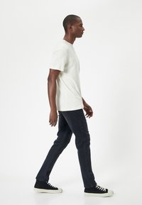 Ljusbeige kortärmad t-shirt, mörk denimjeans och svarta sneakers med vita sulor. Modell som går i en neutral bakgrund.