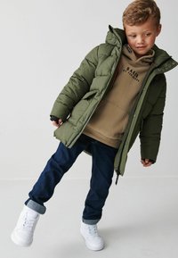 Giacca a vento verde con cappuccio e chiusure a scatto, indossata sopra un felpa marrone. Jeans scuri e sneakers bianche completano l'outfit.