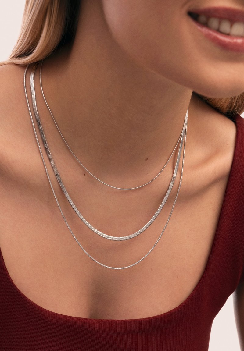 Collana multi-strato in argento con tre strati lisci e curvi di lunghezze diverse, esposta su fondo di una maglietta rossa.