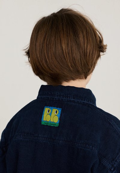 Enfant aux cheveux courts et bruns portant une veste en jean bleu foncé avec un patch logo coloré Bobo Choses dans le dos.