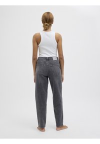 Kvinna med uppsatt hår står barfota, iklädd en vit ärmlös topp och löst sittande mörkgrå jeans mot en enfärgad bakgrund.