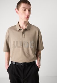 Beige polo shirt med et stort "HUGO"-logo i en lysere nyanse, laget av glattt stoff, parret med sorte bukser og et svart belte.