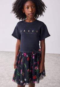 Schwarzes Kurzarm-Top mit goldenem Schriftzug, kombiniert mit einem mehrlagigen, knöchellangen Rock mit floralem Muster in leuchtenden Farben.