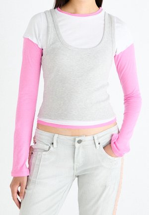 Osoba ubrana w różową i białą warstwową bluzę z długim rękawem pod bezrękawnik w szarym crop topie, do tego jasnoszare dżinsy z różowymi paskami po bokach.