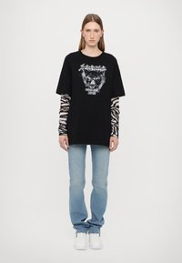 Svart oversized t-skjorte med grafisk design av en panter og tekst. Laget med langermet topp i zebra-mønster. Lyseblå jeans og hvite sneakers.