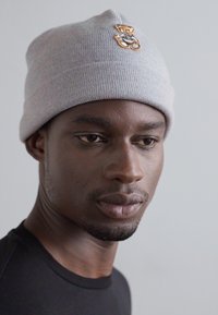 Halli halliga beanie, millel on volditud äär ja mille esiküljel on tikitud karu disain. Sile tekstuur ja tihe sobivus.