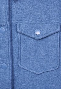 Blauwe textuurstof met een grote voorzak en cirkelvormige blauwe knoopsluiting, met een gladde afwerking en subtiel patroon.