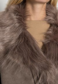 Cuello de piel sintética marrón sobre un abrigo de tela texturizada marrón, superpuesto a una blusa de color beige claro. Textura suave de alta calidad con una apariencia esponjosa.
