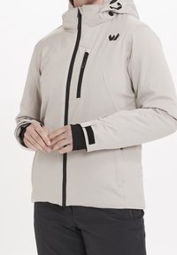 Veste à capuche gris clair avec fermeture éclair noire, poche poitrine et sangles aux poignets, portée par une personne avec les mains jointes devant.