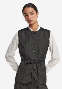 Svart herringbone-väst med kragknäppning, framtill knappar och en knytning i midjan, över en vit blus med långa ärmar.