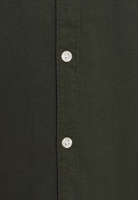 Tummanvihreä, ribbineulottu kangaspaita, jossa valkoiset muovinapit rintalaskoksen varrella; näkyvä tikkauskoriste korostaa pintaa.