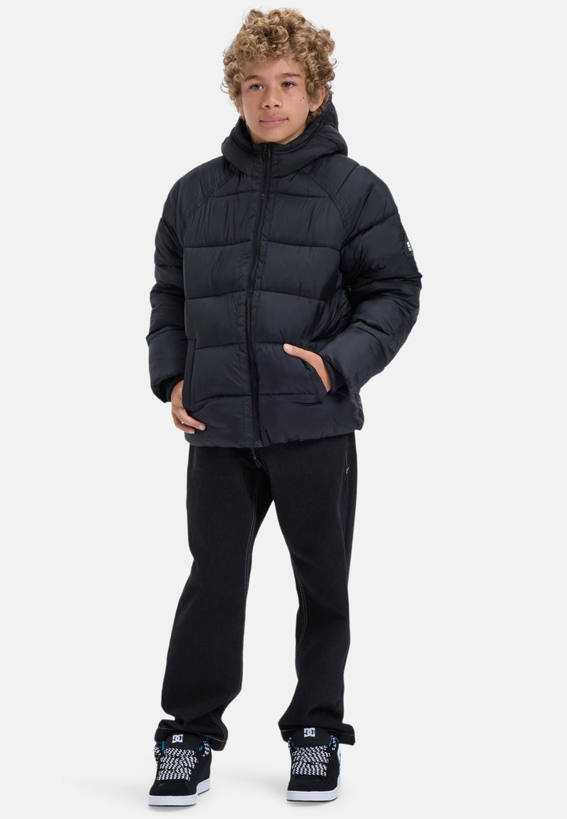 Ung person med lockigt hår som bär en svart puffjacka, svarta byxor och svarta sneakers med mönstrade snören, stående med händerna i fickorna.