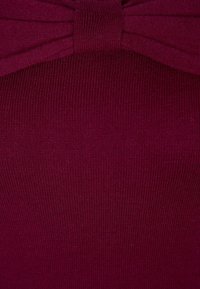 Tessuto a maglia bordeaux con una trama morbida, caratterizzato da un prominente dettaglio a fiocco al centro superiore, creando un design strutturato.