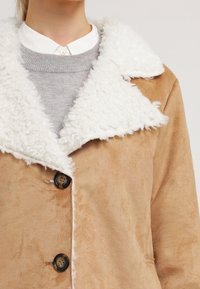 Beige faux-suede jacka med en vit, fluffig krage. Har två stora knappar och en strukturerad yta. Lager på lager med en grå tröja under.