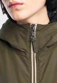 Veste à capuche vert olive avec une fermeture éclair à l'avant en beige contrastant. Dotée d'un col haut et d'un tissu texturé pour l'isolation.