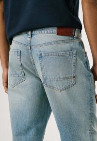 Helles Blau, Rückansicht von Jeans mit zwei Pattentaschen und einem braunen Leder-Logo-Tag am Bund. Weiche Textur, lässige Passform.