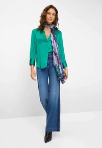 Groene satijnen blouse met knoopdetails, gecombineerd met wijde blauwe spijkerbroek en een gestreepte paarse sjaal, afgemaakt met zwarte puntige laarzen.