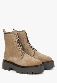 Beżowe zamszowe botki za kostkę o teksturowanym wykończeniu, z delikatnymi perforacjami, sznurowanym przodem oraz masywną ciemną podeszwą z rowkami.
