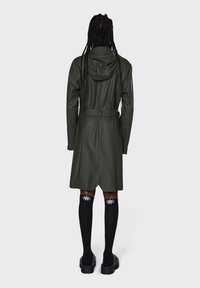 Ελαιόχρωμο αδιάβροχο παλτό με κουκούλα, ζώνη στη μέση και μεσαίου μήκους σχέδιο. Συνδυάζεται με μαύρες κάλτσες μέχρι το γόνατο και χοντρά παπούτσια.